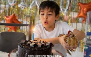Young boy blowing out candles on a chocolate birthday cake while holding a chicken, with balloons in the background.