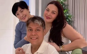 Three people posing indoors: a man sits in front while two women stand behind him, all smiling at the camera.