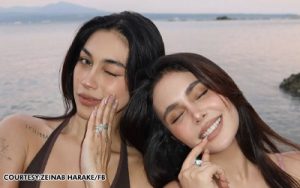 Two women at the seaside with eyes closed, smiling softly, resting their hands on their faces in swimsuits by the water.