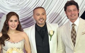 Three people in formal attire posing for a photo at a event: a woman in a strapless gown, a man in a white suit with a black vest, and another man in a light beige suit.
