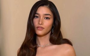 Portrait of a woman with long dark hair wearing a necklace and earrings, gazing at the camera against a beige backdrop.