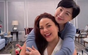 Two women smiling and hugging in a stylish living room, with lamps and chairs in the background.