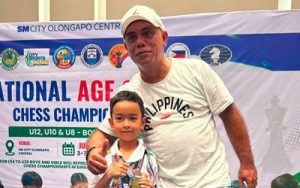 Man and boy pose on a stage at a national chess championship, the boy holding a trophy in front of a banner with event logos.