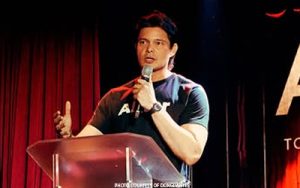 Man in a dark T-shirt speaks into a microphone at a lectern on a stage with red curtains, gesturing with his left hand.