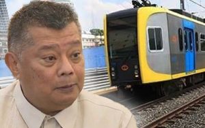 Close-up of a man in a beige jacket with a yellow-and-blue train in the background at a railway station.] ,